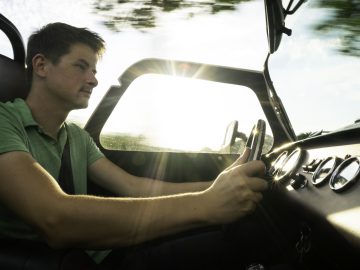 Een jonge man die in een Caterham Super Seven 1600 cabriolet rijdt, geconcentreerd op de weg, terwijl het zonlicht door de bomen stroomt.