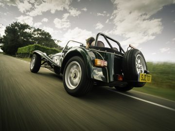 Een persoon die een klassieke Caterham Super Seven 1600 converteerbare sportwagen bestuurt op een weg, bewegend weergegeven met een onscherpe achtergrond.