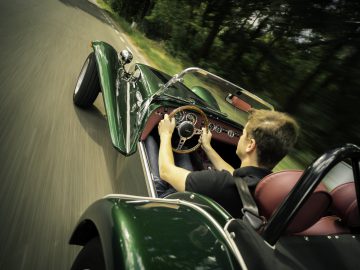 Een man die een klassieke groene Caterham Super Seven 1600 converteerbare sportwagen bestuurt op een weg, gezien vanuit het perspectief van de passagier.