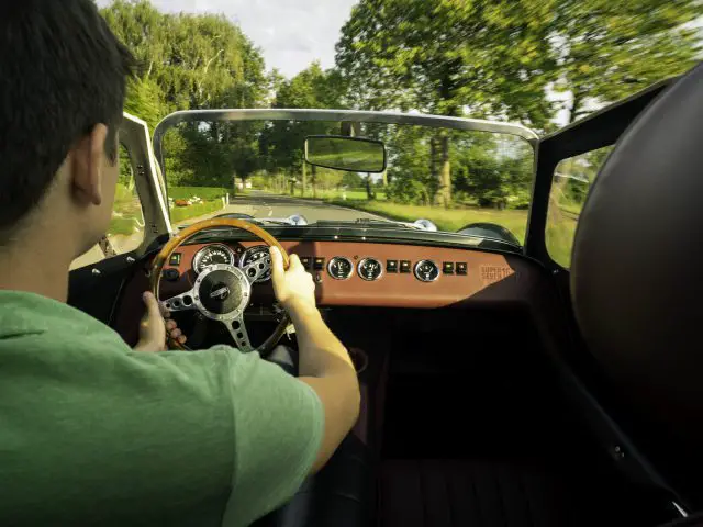 Man bestuurt een Caterham Super Seven 1600 cabriolet met een houten dashboard, gezien vanaf de passagiersstoel, op een zonnige weg omgeven door bomen.