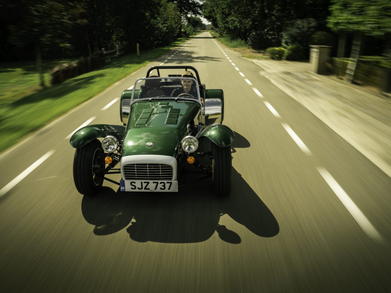 Een groene Caterham Super Seven 1600 cabriolet die met de kap naar beneden over een met bomen omzoomde landweg raast, met een bestuurder en één passagier aan boord.