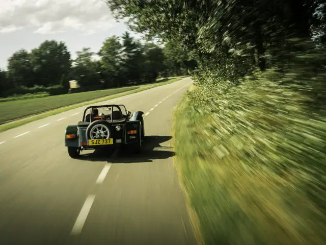 Een klassieke Caterham Super Seven 1600 cabriolet die op een zonnige dag over een weelderige, met bomen omzoomde weg rijdt.