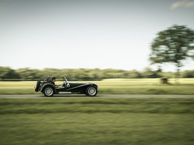 Een klassieke Caterham Super Seven 1600 cabriolet die over een schilderachtige landweg rijdt met groene velden en een enkele boom op de achtergrond.