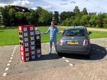 Man die op een parkeerplaats tussen een stapel producten in dozen en een kleine Fiat 500 staat en glimlachend naar de dozen gebaart.