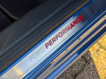 Close-up van een metalen Ford Focus ST-instaplijsten met het opschrift "Ford Performance" met een blauwe en rode streep, gezien vanuit een hoek.