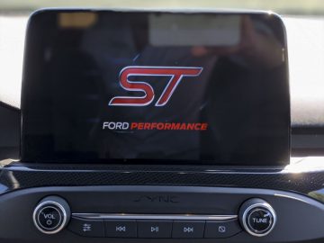 Het digitale display van een auto met het Ford Focus ST-logo, gemonteerd boven het dashboard met zichtbare bedieningsknoppen eronder.