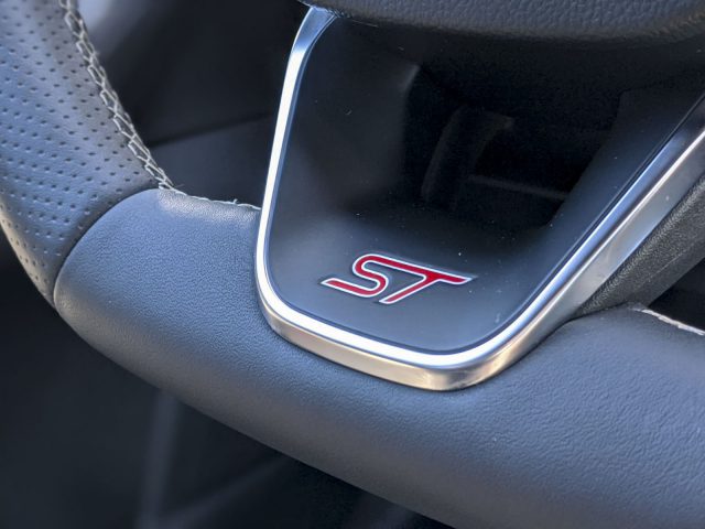 Close-up van het stuur van een Ford Focus ST met het "ST"-logo op een zilveren badge, tegen een zwartleren achtergrond.