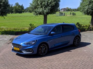 Een blauwe Ford Focus ST geparkeerd op een parkeerplaats met groene velden en bomen op de achtergrond op een zonnige dag.