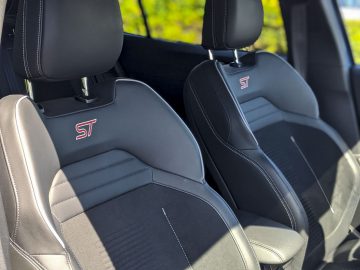 Binnenaanzicht van een Ford Focus ST met twee sportieve stoelen met het "ST"-logo op de hoofdsteunen, voorzien van zwarte en grijze bekleding.