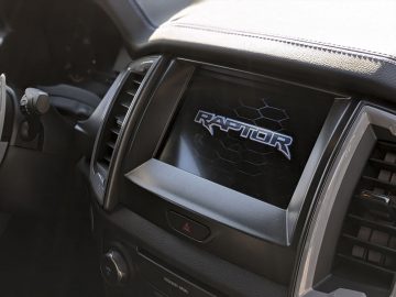 Dashboard van de auto met een 'Ford Ranger Raptor'-logo in een gestileerd frame, wat het robuuste model van de auto benadrukt.
