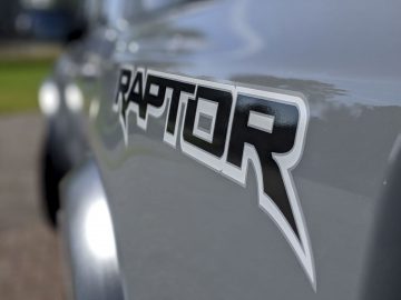 Close-up van het "raptor"-logo op de zijkant van een grijze Ford Ranger, met een zachte focus op de achtergrond.