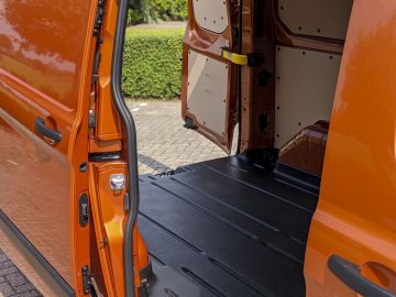 Open achterdeur van een oranje Ford Transit Custom-busje met een lege laadruimte met zwarte vloer, gezien vanaf de buitenkant.