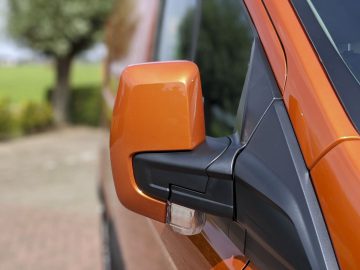 Close-up van de zijspiegel van een Ford Transit Custom met een duidelijke focus op de behuizing, tegen een onscherpe achtergrond van een oprit en bomen.