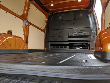 Binnenaanzicht van een lege Ford Transit Custom bestelwagen met oranje en grijze muren, met de geribbelde metalen vloer en wielkasten.