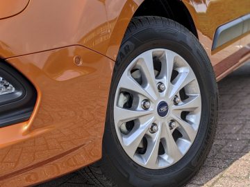 Close-up van het voorwiel en de band van een oranje Ford Transit Custom, met een zilveren wieldop met het Ford-logo, geparkeerd op een bakstenen oppervlak.