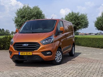 Oranje Ford Transit Custom-busje geparkeerd op een verhard terrein met gras en lucht op de achtergrond.