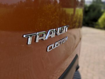 Close-up van een 'Ford Transit Custom'-embleem op de zijkant van een oranje voertuig, met een wazige groene achtergrond.