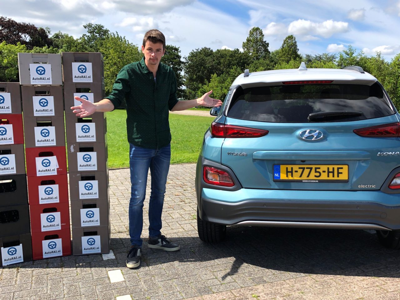 Man gebaart naar talloze gestapelde dozen met verschillende logo's naast een blauwe elektrische auto van Hyundai Kona in een zonnige buitenomgeving.