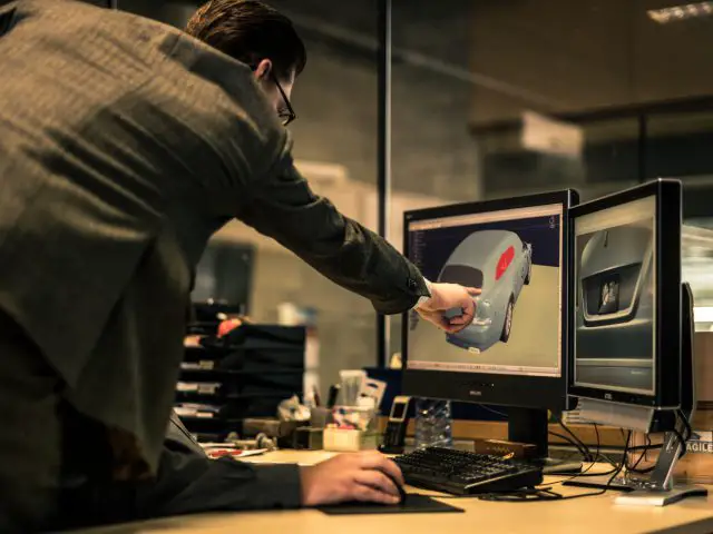 Een man in pak wijst naar een computerscherm met een 3D-model van de Rolls-Royce Silver Spectre Shooting Brake in een kantooromgeving.