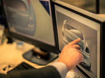 De vinger van een persoon die het stuur van een Rolls-Royce Silver Spectre Shooting Brake aanraakt op een computerscherm, wat een ontwerp of functie in een zakelijke kantooromgeving aangeeft.