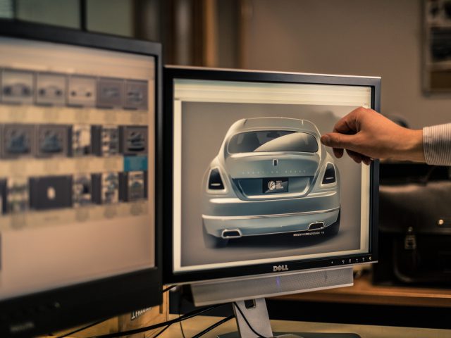 Persoon die naar een computerscherm wijst met een 3D-model van een Rolls-Royce Silver Spectre Shooting Brake in een kantooromgeving.