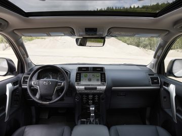 Binnenaanzicht van een Toyota Land Cruiser met het stuur, het dashboard en de stoelen met een panoramisch schuifdak open, met uitzicht op een grindgebied buiten.