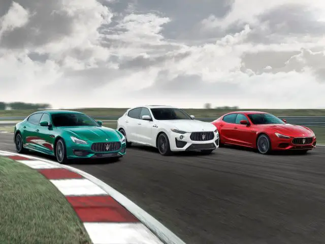 Drie Maserati Ghibli-auto's racen op een circuit; een groene, een witte en een rode auto, elk rijdend op een bewolkte dag.