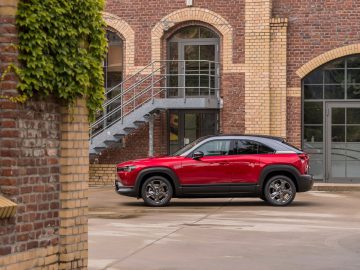 Een rode Mazda MX-30 geparkeerd tussen bakstenen gebouwen met boogramen en een metalen trap.
