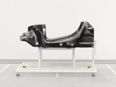Een zwart McLaren-autochassisonderdeel weergegeven op een witmetalen standaard in een minimalistische industriële ruimte.