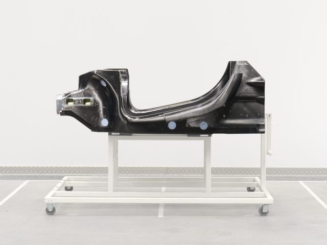 Een zwart McLaren-autochassisonderdeel weergegeven op een witmetalen standaard in een minimalistische industriële ruimte.