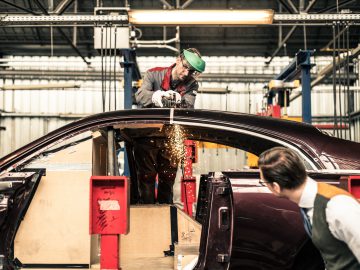 Een arbeider in beschermende uitrusting gebruikt elektrisch gereedschap op een Rolls-Royce Silver Spectre Shooting Brake-carrosserie in een industriële werkplaats, terwijl een andere persoon toekijkt.