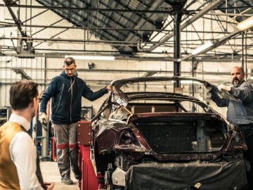 Twee mannen werken in een werkplaats aan een Rolls-Royce Silver Spectre Shooting Brake, terwijl een andere man hen observeert. Ze staan in een ruime garage met gereedschap en apparatuur.