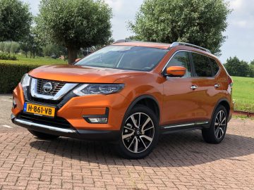 Een feloranje Nissan X-Trail SUV geparkeerd op een verhard terrein met groen gras op de achtergrond op een zonnige dag.