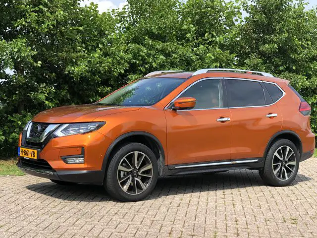 Een feloranje Nissan X-Trail SUV geparkeerd buiten met bomen op de achtergrond op een zonnige dag.