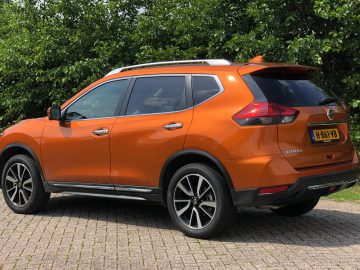 Nissan X-Trail SUV geparkeerd buiten naast groene struiken onder een heldere hemel.