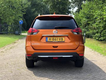 Achteraanzicht van een oranje Nissan X-Trail geparkeerd op een straat met bomen op de achtergrond op een zonnige dag.