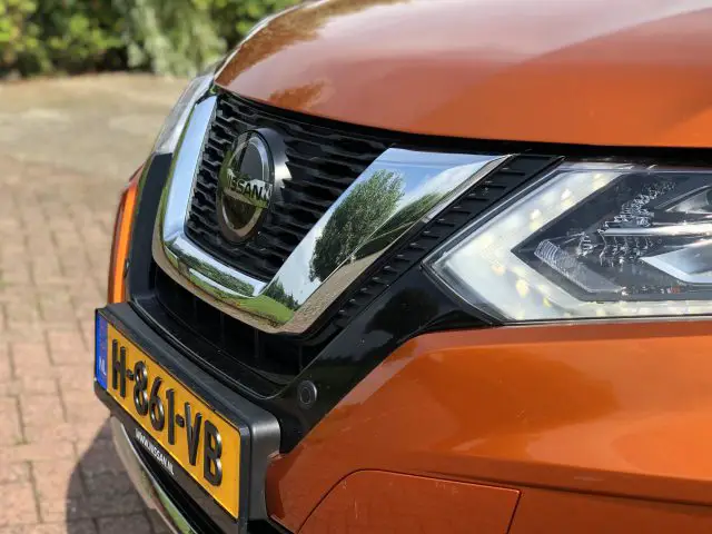 Close-up van de voorkant van een oranje Nissan X-Trail-voertuig, met de nadruk op de koplamp, grille en badge met een wazige groene achtergrond.