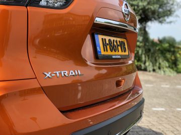 Close-up van de achterkant van een oranje Nissan X-Trail met zichtbare kentekenplaat, buiten geparkeerd.