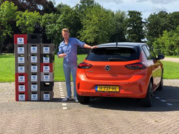 Een man stond naast een rode Opel Corsa geparkeerd voor een display met meerdere borden, wijzend naar het display.