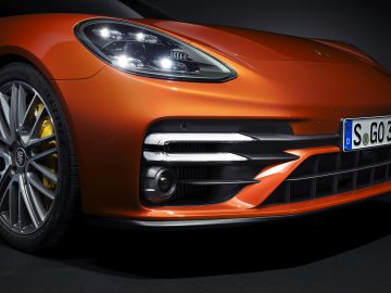 Close-up van het voorste gedeelte van een oranje Porsche Panamera-sportwagen met gedetailleerd zicht op de koplamp, grille en kentekenplaat.
