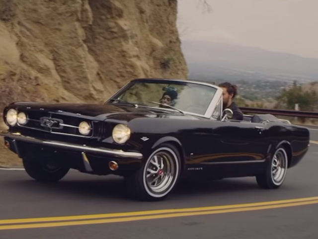 Een zwarte Ford Mustang cabriolet uit 1965 met Lisa Bonet en Jason Momoa die over een bochtige bergweg rijden.