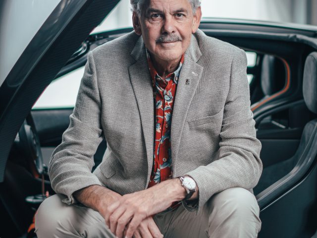 Oudere man met een snor, zittend op de drempel van de Gordon Murray Automotive T.50 auto, gekleed in een grijze blazer, overhemd met patroon en beige broek, glimlachend naar de camera.