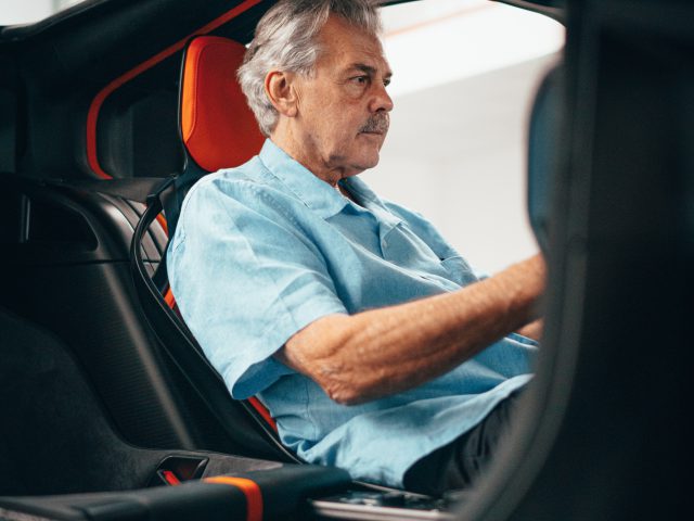 Oudere man met grijs haar zit in een Gordon Murray Automotive T.50, gefocust op autorijden, bekeken door het autoraam.