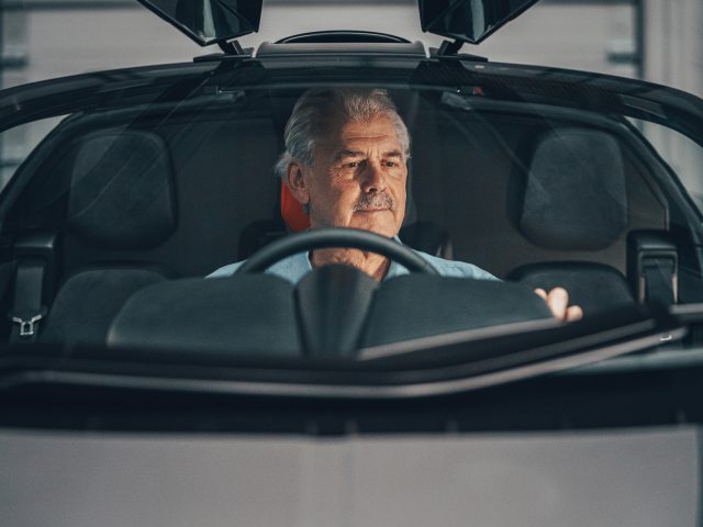 Senior man met een snor die in een Gordon Murray Automotive T.50 rijdt, gezien vanaf de achterklep, en blijk geeft van focus en vastberadenheid.