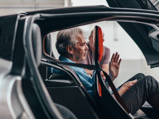 Senior man zit op de bestuurdersstoel van een Gordon Murray Automotive T.50 met open vleugeldeuren, gebarend en pratend met een andere persoon die buiten staat.