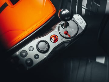 Close-up van de handmatige versnellingspook en bedieningselementen van een gestroomlijnde Gordon Murray Automotive T.50 met gedeeltelijk zicht op een oranje lederen stoel.