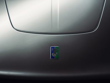 Close-up van de motorkap van een auto met een rechthoekig logo met een gestileerd pauwontwerp tegen een blauwe achtergrond, emblematisch voor de Gordon Murray Automotive T.50.