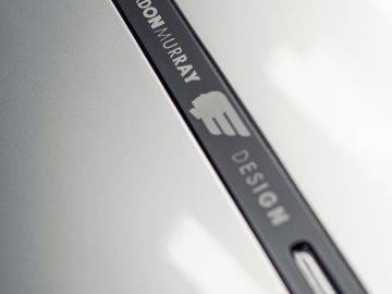 Close-up van een metalen naamplaatje met de gravure "Gordon Murray Automotive T.50" op een strak grijs oppervlak.