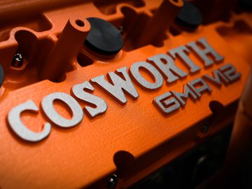 Close-up van een oranje Gordon Murray Automotive T.50-motorkap met gedetailleerde textuur en branding in zilveren letters.