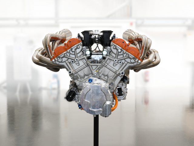 Een Gordon Murray Automotive T.50-motor tentoongesteld op een standaard in een schone, goed verlichte industriële ruimte, met oranje details en complexe leidingen.